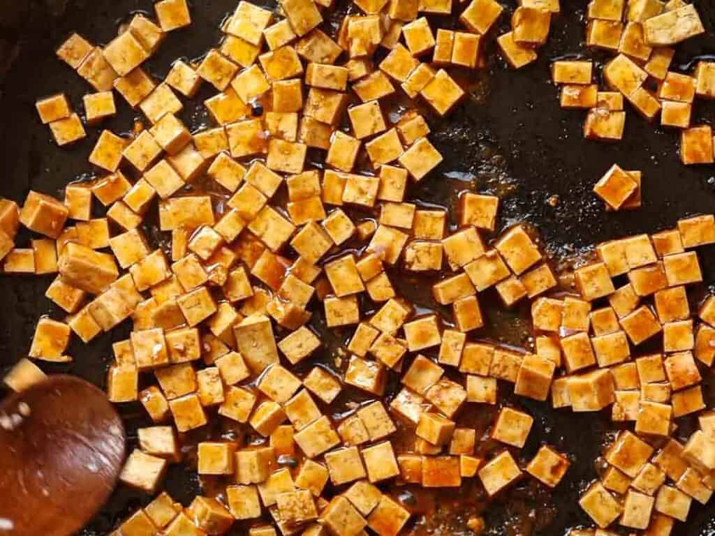 Cooking tofu cubes are scattered in a pan, coated with a brown sauce. A wooden spoon is partially visible on the left side, stirring the mixture. The tofu appears golden and evenly distributed across the pan.